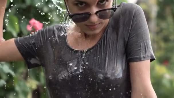 Jeune jolie fille percée dans les lunettes de soleil s'amuser avec pulvérisation d'eau de tuyau dans le jardin. T-shirt de loisirs d'été et humide. Beau corps — Video