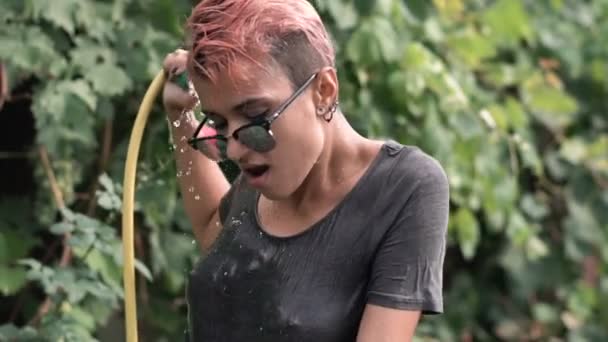 Jeune jolie fille percée dans les lunettes de soleil s'amuser avec pulvérisation d'eau de tuyau dans le jardin. T-shirt de loisirs d'été et humide. Beau corps — Video