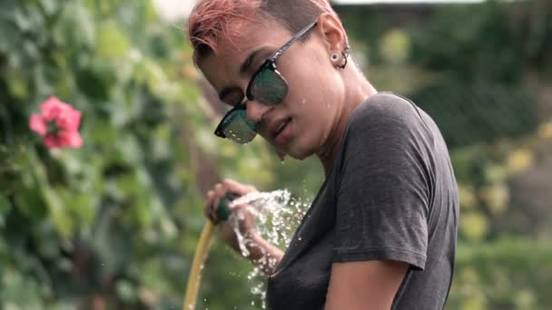 Young Pretty Pierced Girl en gafas de sol divirtiéndose con spray de agua de la manguera en el jardín. Camiseta de verano de ocio y mojada. Hermoso cuerpo — Vídeo de stock