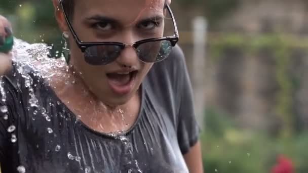 Jeune jolie fille percée dans les lunettes de soleil s'amuser avec pulvérisation d'eau de tuyau dans le jardin. T-shirt de loisirs d'été et humide. Beau corps — Video