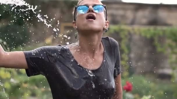 Молодая красотка в солнечных очках, развлекающаяся с брызгами воды из шланга в саду. Летний отдых и мокрая футболка. Тело — стоковое видео