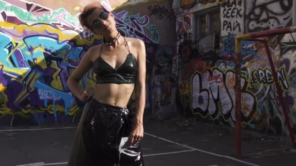 Straßenpunk oder Hipster-Mädchen mit rosa gefärbten Haaren. Frau mit Piercing in Nase, Ohren und ungewöhnlicher Frisur steht mit Dollars in der Hand auf Basketballplatz mit Graffiti-Bildern an Wänden — Stockvideo