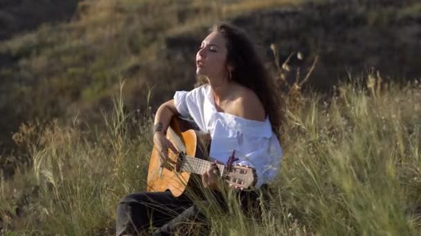 Σγουρά Gipsy μελαχρινή κοπέλα παίζοντας κιθάρα και τραγουδώντας στο πεδίο στη πλαγιά του κόλπου — Αρχείο Βίντεο