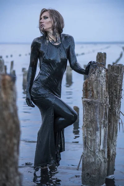 Retrato de la chica rubia caucásica se mantiene untada en un barro negro saludable en el viejo Firth con postes de madera para la producción de sal — Foto de Stock