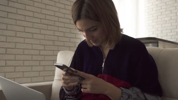 Una chica escribiendo en el teléfono móvil, luego sonriendo y escribiendo en un ordenador portátil. Hembra escribiendo en el teclado. Freelancer chica trabajando desde casa — Vídeo de stock