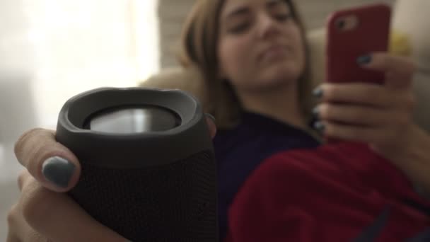 Hermosa chica acostada en el sofá y escuchando música en el teléfono inteligente desde el altavoz inalámbrico, que está sosteniendo en la mano . — Vídeos de Stock