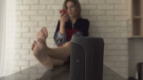 Um alto-falante sem fio preto em pé em uma mesa de cozinha. Menina bonita sentada no fundo encostada à parede movendo a cabeça para o rítmo e ouvir música — Vídeo de Stock