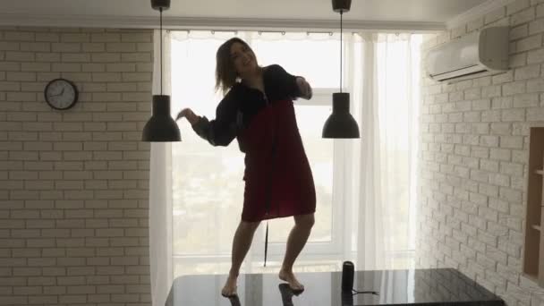 En smuk brunette pige i en rød fløjlskjole danser barfodet på det sorte køkkenbord med en mobiltelefon i hånden og en trådløs højttaler på bordet – Stock-video