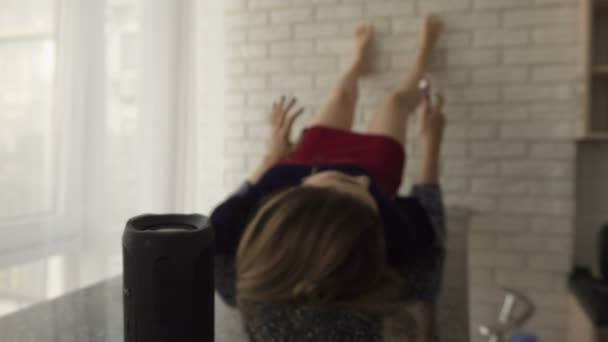 En svart trådlös högtalare stående på ett köksbord. Vacker flicka liggande i bakgrunden på samma tabell med benen upp på väggen och dans till rytm och lyssna på musik — Stockvideo
