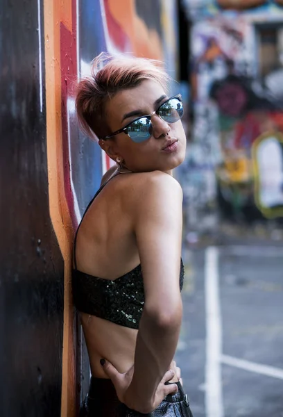 Street punk or hipster girl with pink dyed hair. Woman with piercing in nose, ears tunnels and unusual hairstyle posing at basketball court with graffiti paintings on walls