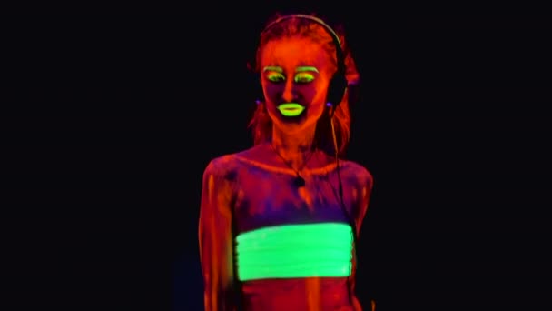 Eine schöne junge trägt Kopfhörer sexy halbnackte Mädchen verführerisch tanzen mit UV-Farbe auf ihrem Körper. hübsche Frau mit glühendem Körper im Schwarzlicht. — Stockvideo