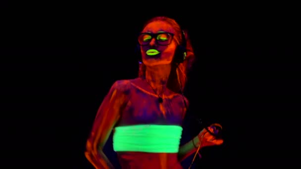 Une belle jeune fille sexy en faux lunettes et écouteurs dansant avec de la peinture éclatante sur son corps à la lumière noire. Jolie femme avec bodyart lumineux dans la lumière de la lampe noire . — Video