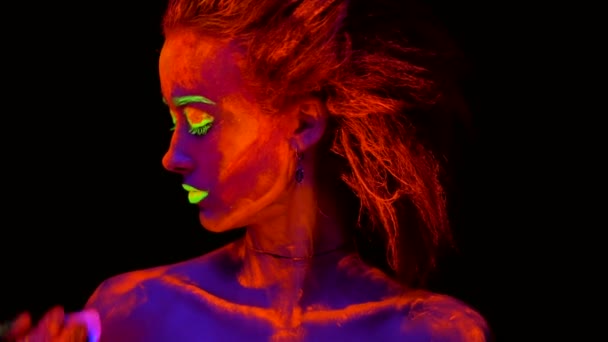 Una hermosa joven sexy con pintura ultravioleta en su cuerpo mirando una mano de un artista con un pincel, pintando su hombro. Mujer bonita con el bodyart brillante en la luz de la lámpara negra . — Vídeo de stock
