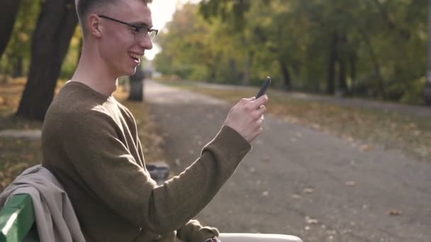 Fiatal Streetstyle-t az ember a termékeinek élvez használ a smartphone a parkban, a kaukázusi fiatalember szemüveg — Stock videók
