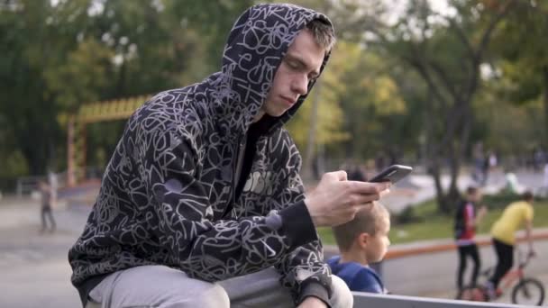 パーカーのフードの彼のスマート フォンを使用しての若いストリート スタイル男のスケートパークのフェンスの上に座って — ストック動画