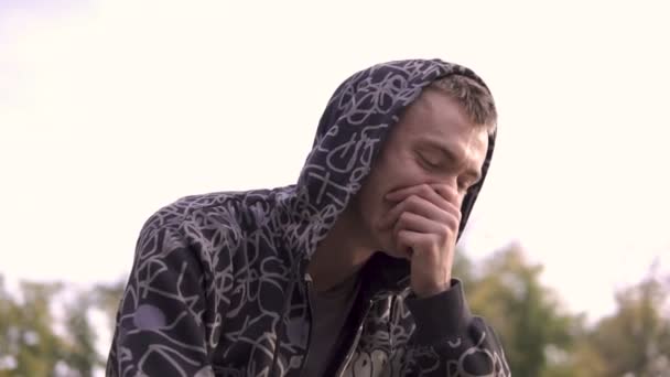 Ung streetstyle mand i hættetrøje Laughting mens du bruger sin smartphone i hætte, Sidder på hegnet af skatepark – Stock-video