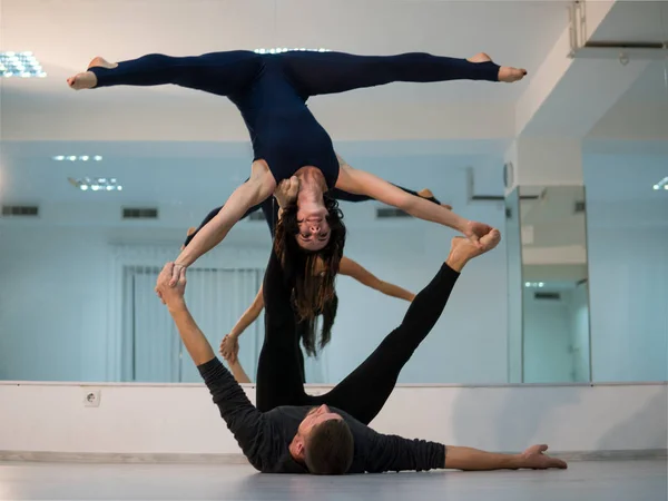 Jovem casal atlético em ternos escuros praticando acroyoga em estúdio com espelhos. Balanceamento em par — Fotografia de Stock