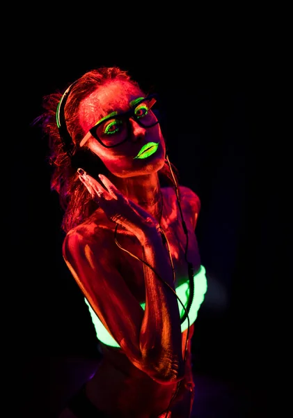 Retrato de una hermosa joven sexy en auriculares con pintura ultravioleta en su cuerpo. Mujer bonita con el bodyart brillante en la luz de la lámpara negra — Foto de Stock