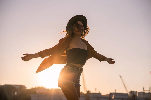 Lockigt kastanj-haired sexig tjej i jeansshorts och hatt snurrar på havet port broadway i solnedgången, i industriella stadsmiljö — Stockfoto