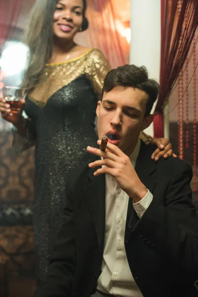 YOung stylish caucasian Man in black suit smoking cigar and drinking whiskey at night in a company with pretty african girl