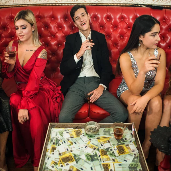 Group of young multi-ethnic friends relaxing in shisha club-bar in rich interior. Man with cigar counting money in the club
