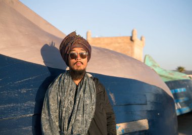 Genç Asyalı turist sakallı adam Dreadlocks ve kalış kafasına eşarp duymak mavi balıkçı tekne Essaouira, Fas akşam günbatımı zaman