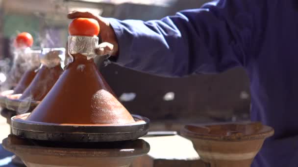 传统的摩洛哥塔吉食品烹饪在塔金锅的火与烟雾和西红柿在上面。当地厨师的手正在打开沸腾的菜, 并在阳光明媚的天气释放烟雾 — 图库视频影像