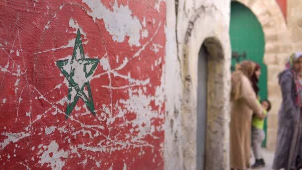 Bandera de Marruecos con una bandera verde estrella de Marruecos en la pared de la calle en Essaouira Medina. La gente local está pasando por una calle borrosa — Vídeos de Stock