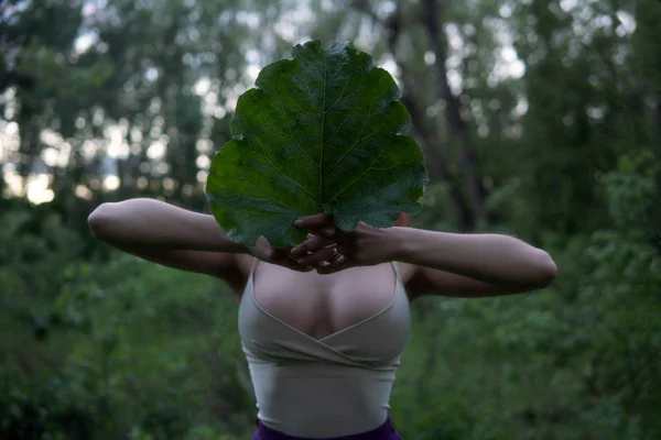 Junge Frau mit großen Titten versteckt ihr Gesicht hinter einem großen Klettenblatt im Wald in der geheimnisvollen Dämmerung — Stockfoto