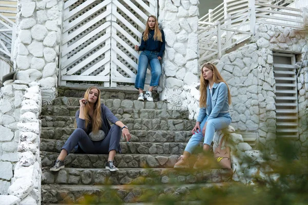 Grupo de tres jóvenes elegantes chicas bonitas en trajes de traje de pantalón sentado en una escalera cerca de la construcción blanca —  Fotos de Stock