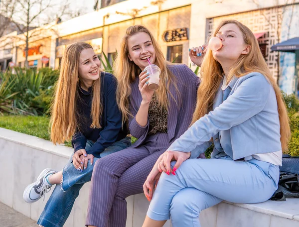 Three girls friends having fun on evening broadway. Eating ice cream, drinking ice fresh milk shake and chewing bubble gum