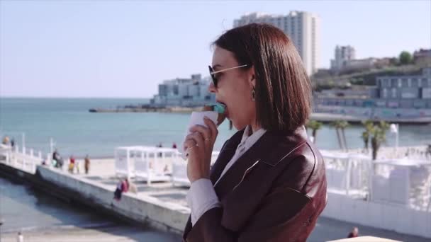 Adorabile ragazza bruna elegante che cammina in banchina vicino al mare e mangia gelato nella giornata di sole — Video Stock