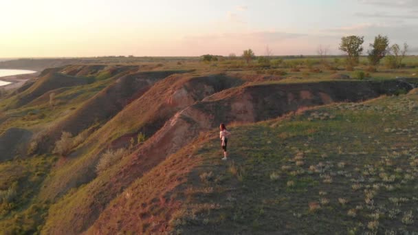 Tiro aéreo de jovem mulher esportiva fazendo exercícios no egde de encosta perto do lago ou liman durante o belo nascer do sol ou pôr do sol — Vídeo de Stock