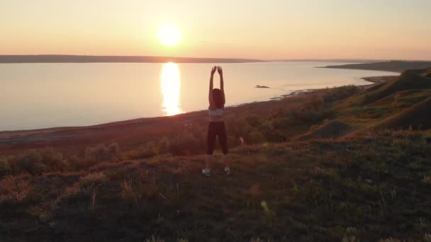 美しい日の出や日没の間に湖やリマンの近くの斜面の例で演習を行う若いスポーティな女性の空中ショット — ストック動画