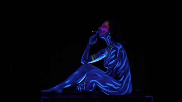 Trupul complet al fetei tinere Adorabile dezbrăcate sexy, care fumează în lumină UV pe fundal negru. Negru lumina stralucitoare vopsea albastru bodyart avatar cosplay doamnă cu fata luminoasa vopsita — Videoclip de stoc