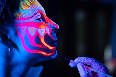 Ultraviyole siyah ışık parlayan bodyart genç kadınlar yüzünde işleme. Soğuk mavi ışıkta pembe ve mor boyalar