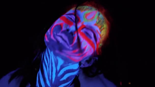 Крупним планом портрет молодої чарівної дівчини в ультрафіолетовому світлі на чорному тлі. Чорне світло світиться фарбою тіло мистецтва аватар косплей леді з пофарбованим емоційним обличчям — стокове відео