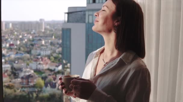 Aantrekkelijke brunette vrouw genieten van haar aromatische koffie en kijken in het venster van hoge vloer op de avond huisjes — Stockvideo