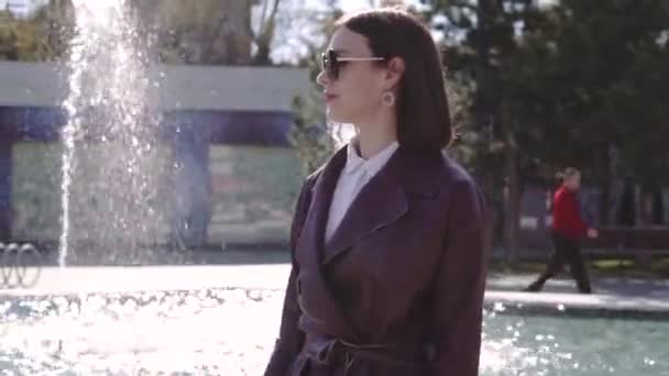 Wanita cantik mewah dalam gaun jaket burgundy berjalan di dekat air mancur — Stok Video