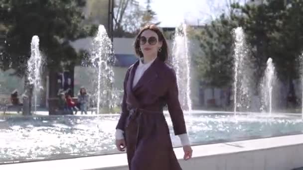 Luxury beautiful woman in burgundy jacket dress walking near the fountain — Stock Video