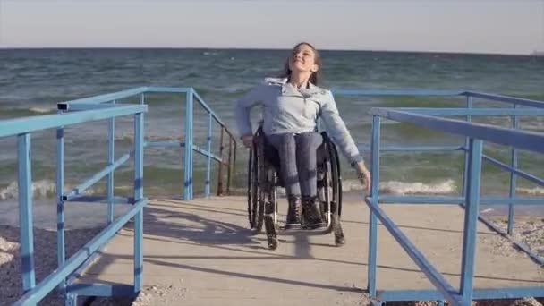 Joven mujer discapacitada en silla de ruedas que se mueve por la rampa para personas con discapacidad cerca del mar en primavera u otoño — Vídeos de Stock