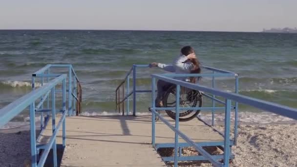 Νεαρή γυναίκα με ειδικές ανάγκες σε αναπηρικό αμαξίδιο που κινείται από τη ράμπα για άτομα με αναπηρίες κοντά στη θάλασσα την άνοιξη ή το φθινόπωρο — Αρχείο Βίντεο