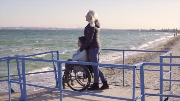 Οικογενειακή αναψυχή. Νεαρή γυναίκα με αναπηρία σε αναπηρικό καροτσάκι με τη μητέρα της να περπατάει κοντά στη θάλασσα στη ράμπα για άτομα με αναπηρίες — Αρχείο Βίντεο