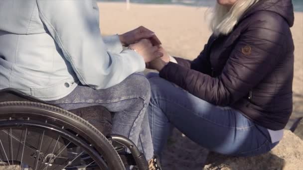 Familie vrije tijd. Jonge gehandicapte gehandicapte vrouw in rolstoel sprekende met haar moeder zittend in de buurt van de zee en houden elkaars handen — Stockvideo