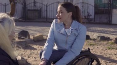 Aile eğlencesi. Tekerlekli sandalyedeki genç engelli kadın, annesiyle deniz kenarında otururken birbirlerinin elleriyle konuşuyor