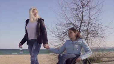 Aile eğlencesi. Tekerlekli sandalyedeki genç engelli kadın, annesi yle birlikte deniz kenarında yürürken, konuşup eğleniyor
