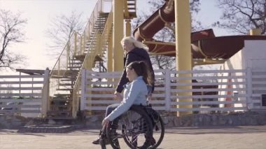 Aile eğlencesi. Tekerlekli sandalyedeki genç engelli kadın, annesi yle birlikte deniz kenarında yürürken, konuşup eğleniyor
