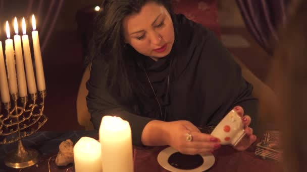 Fortune teller vrouw in donkere kamer met veel kaarsen geeft een uitleg van de toekomst en voorspelling van het lot aan een klant met koffie kopje dik sediment — Stockvideo