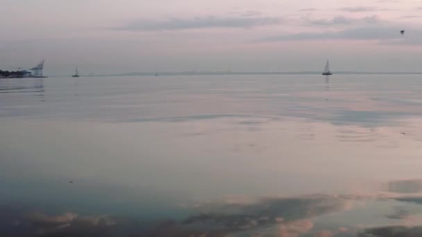 Спокойная утренняя панорама поверхности морской воды. Облака отражаются в зеркале воды во время восхода или захода солнца. Маленькая лодка с парусом на горизонте — стоковое видео
