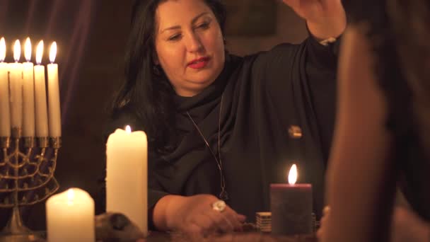Женщина-гадалка в темной комнате с множеством свечей дает объяснение будущего и предсказание судьбы клиенту с помощью золотого обручального кольца — стоковое видео